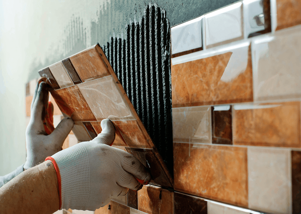 Person installing brown and beige ceramic tiles on a wall with adhesive.